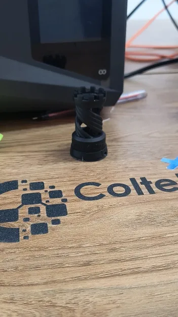 Pieza negra con dentado helicoidal impresa en filamento, sobre la mesa del taller junto al logotipo de Coltek Desing.